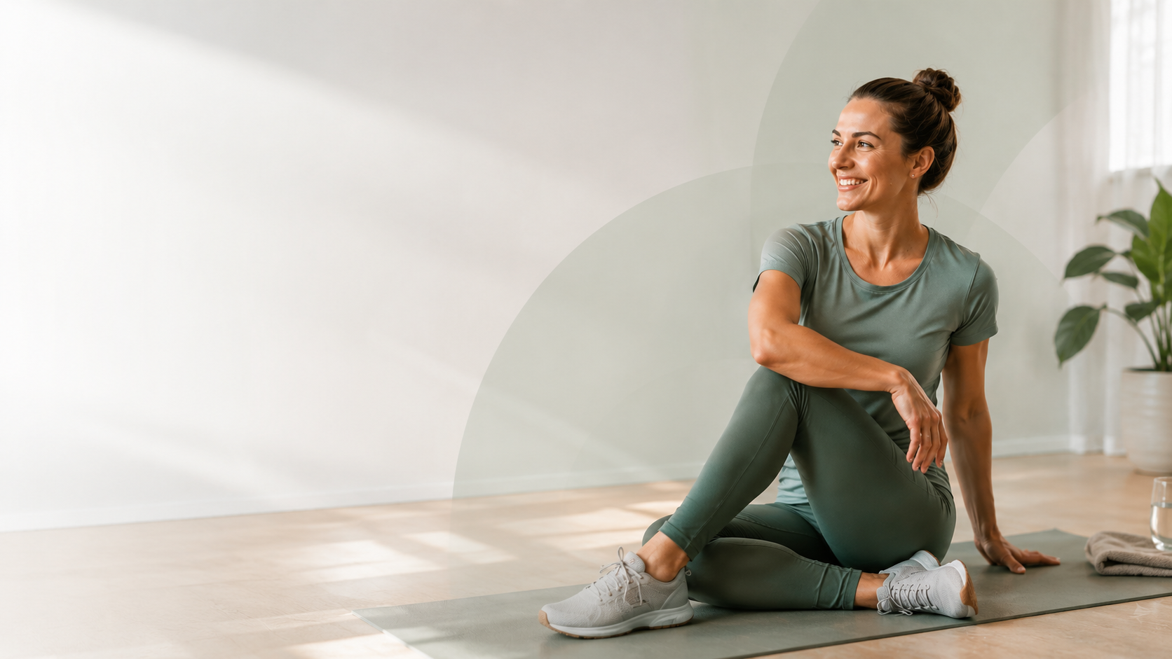 Frau sitzt entspannt lächelnd auf einer Yogamatte in einem hellen Raum zu Hause